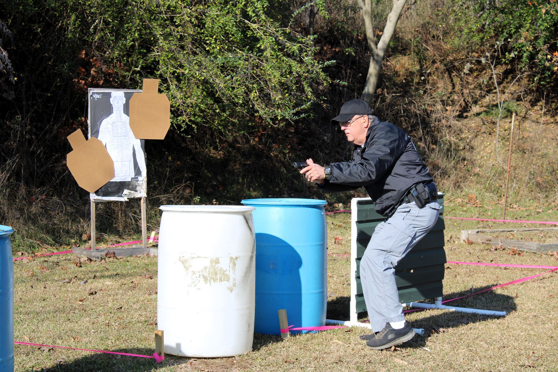 A person aims a handgun at targets on a shooting range, standing between blue and white barrels outdoors.