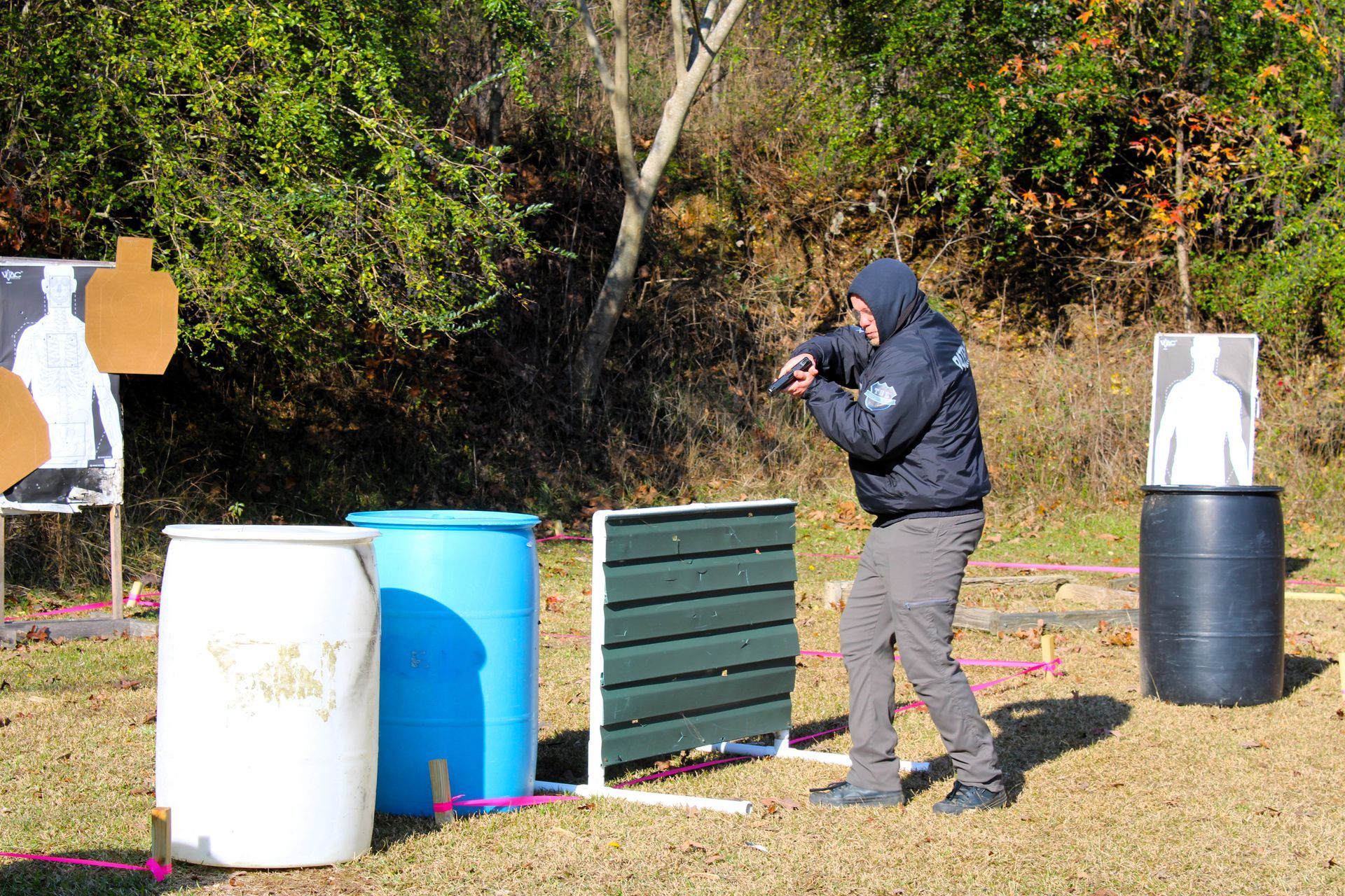 A person in a hooded jacket practicing target shooting at an outdoor range with various barrels and paper targets.