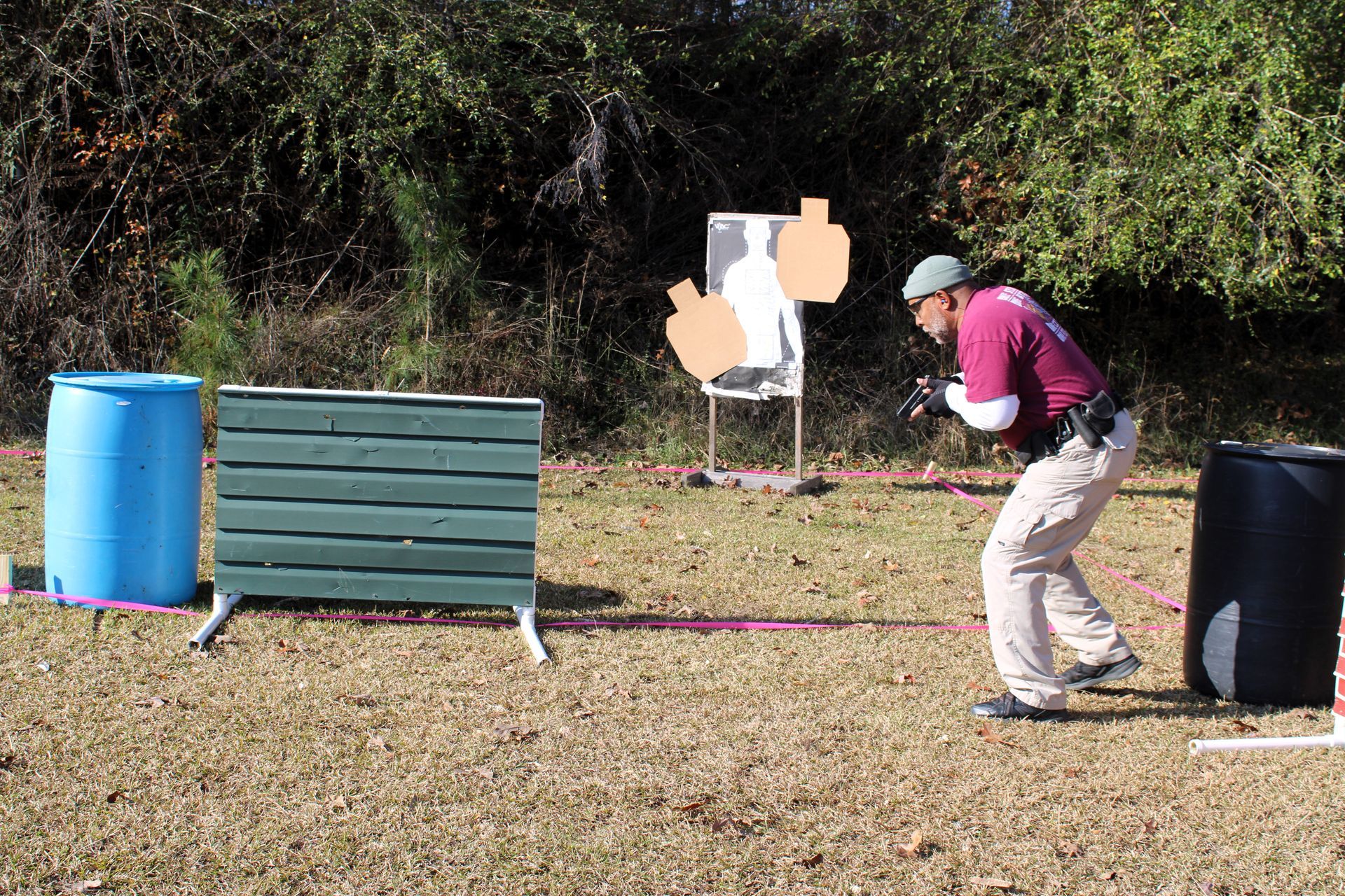 A shooter in a maroon shirt crouches near barrels and a wooden barrier on a grass range while aiming at paper targets.