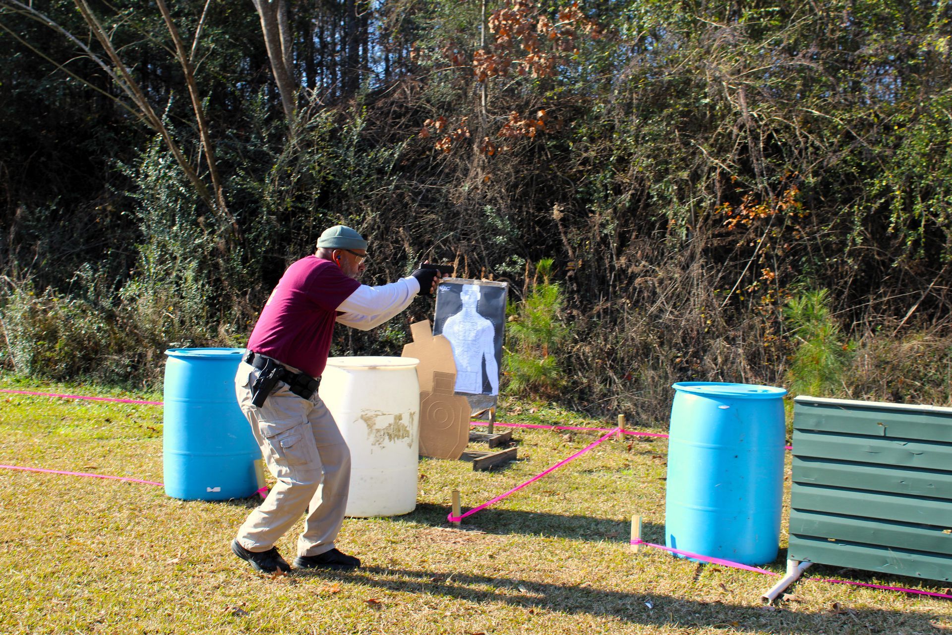 A person in a maroon shirt and tan pants practicing at an outdoor shooting range with blue barrels and a target.