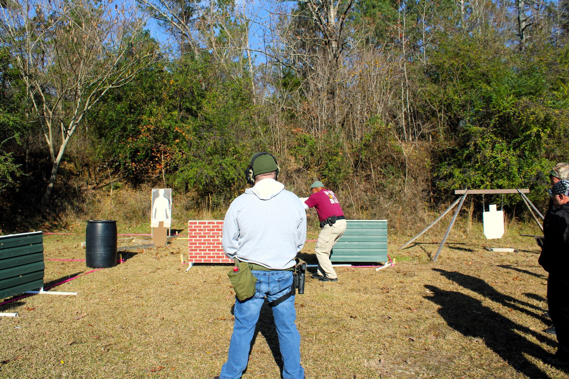 Two people practicing at an outdoor shooting range with paper targets and training barriers in a wooded area.