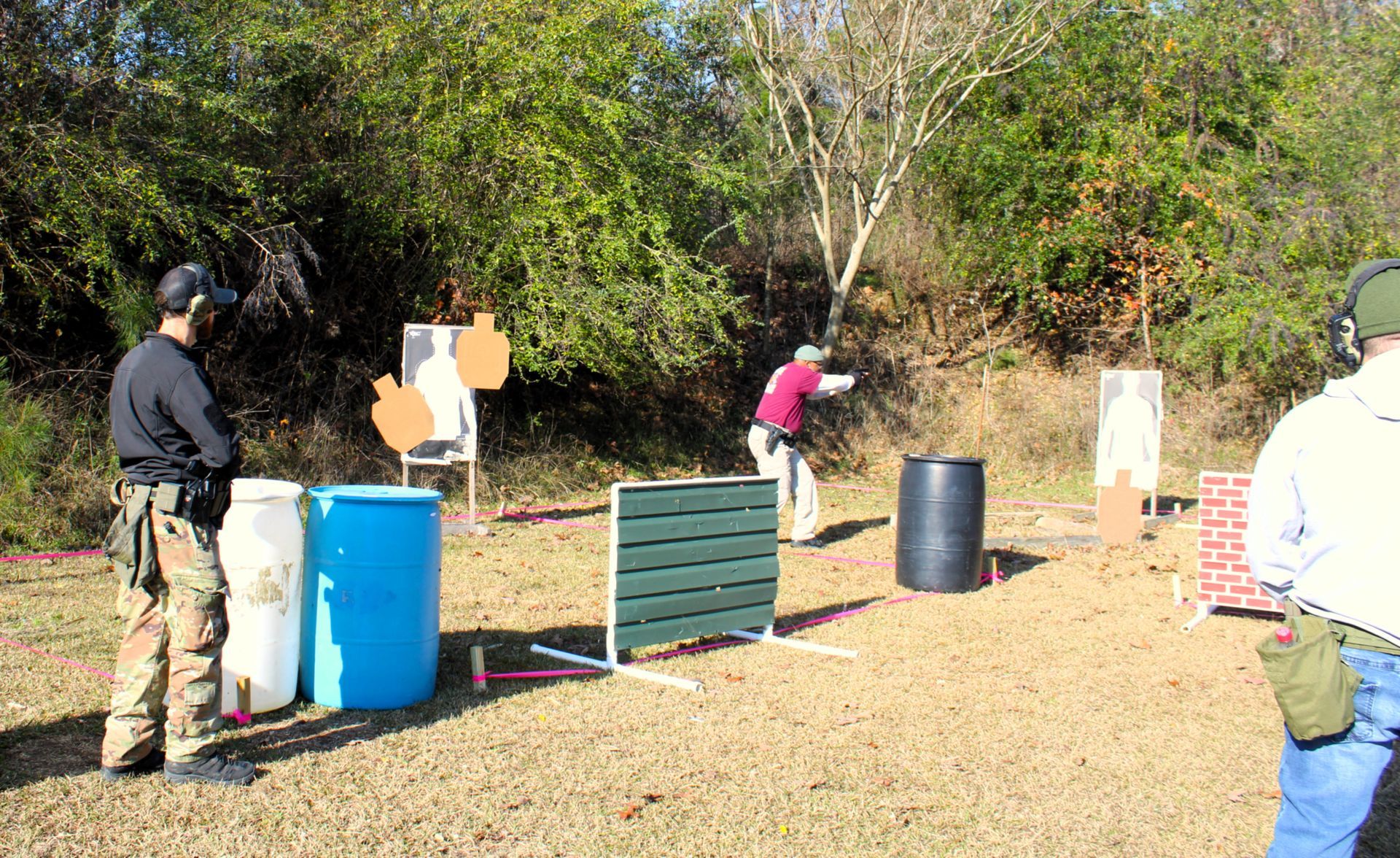Participants at a shooting range practice target shooting with firearms near obstacles and targets in an outdoor field.