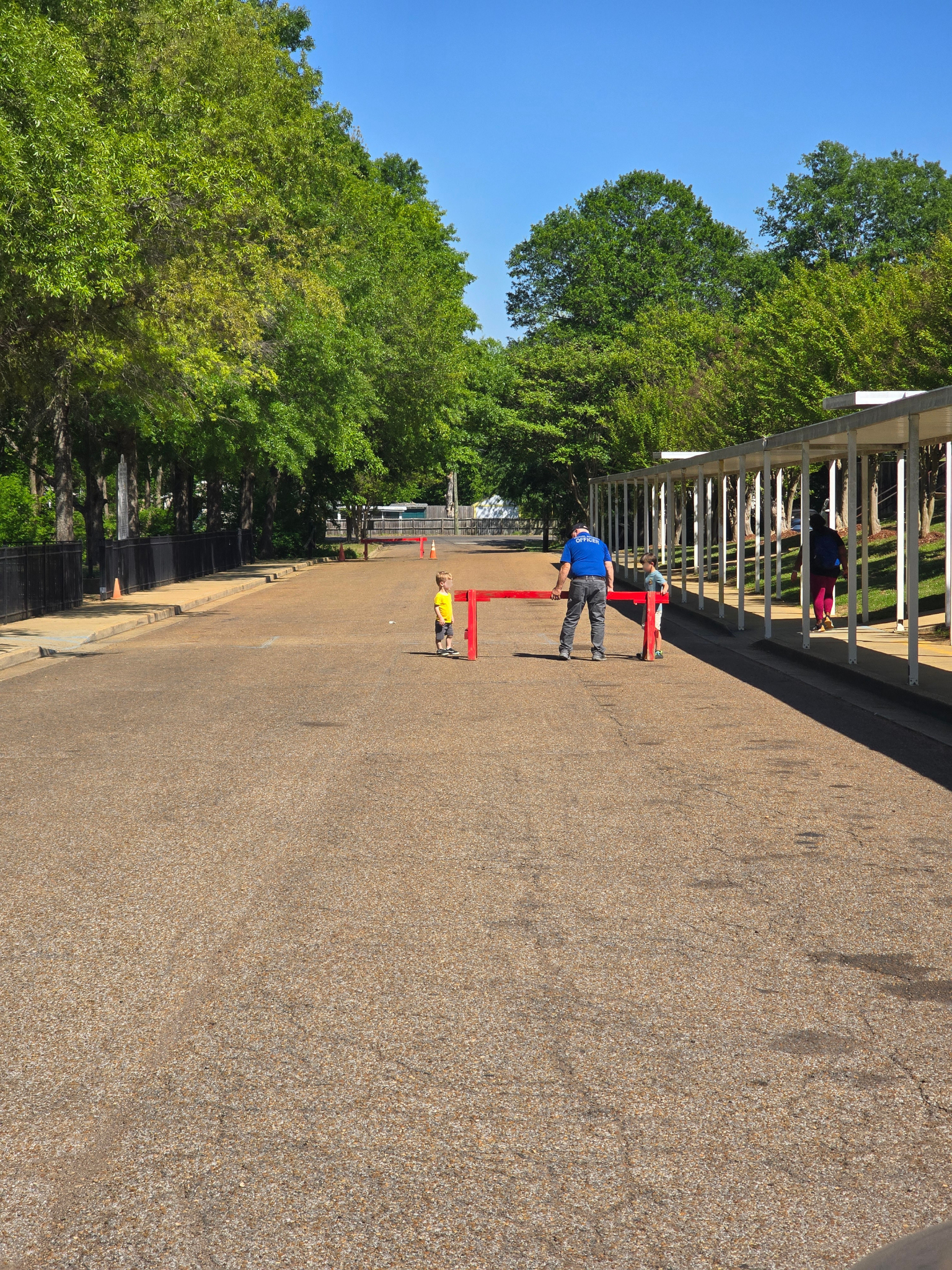 Two people stand near a red barrier on a gravel path lined with trees and a covered walkway on a sunny day.