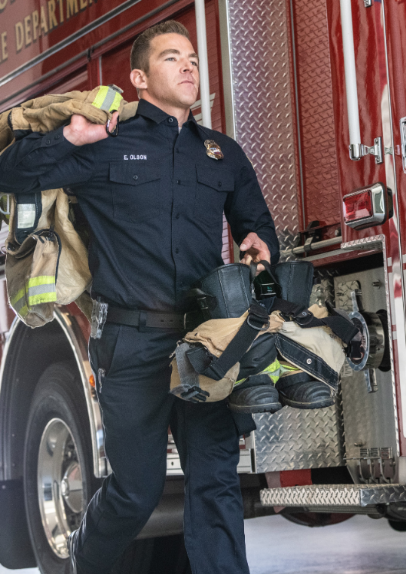 A firefighter in a navy uniform walks past a red fire truck, carrying bunker gear and boots.