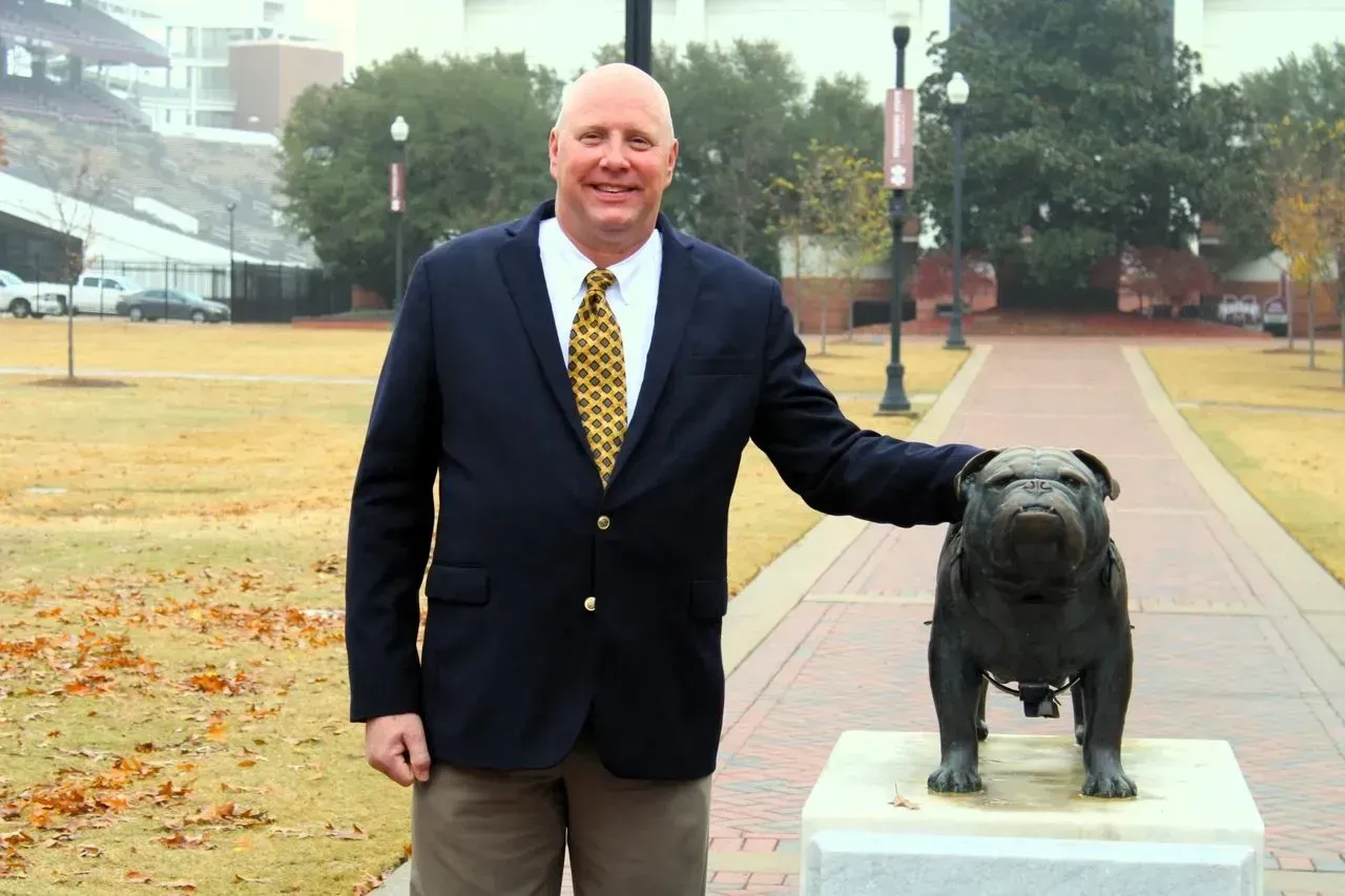A man in a navy blazer and patterned tie stands on a path, resting his hand on a bronze statue of a bulldog.