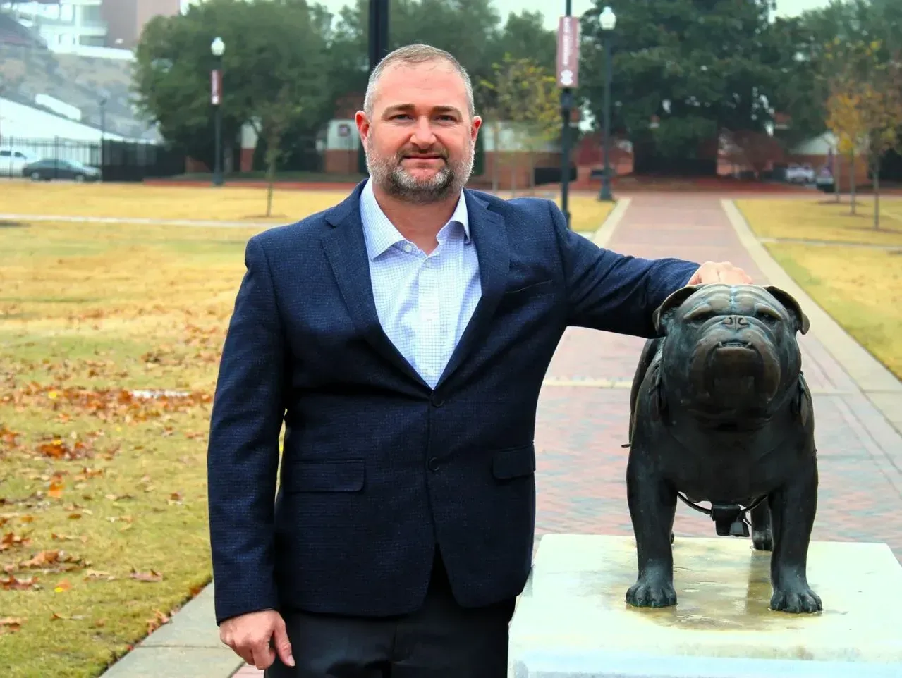 A man in a navy blazer stands outdoors on a brick path, resting his hand on a bronze bulldog statue.