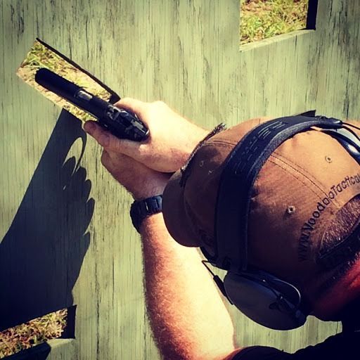 A person wearing ear protection and a cap aims a handgun through a rectangular cutout in a wooden wall.