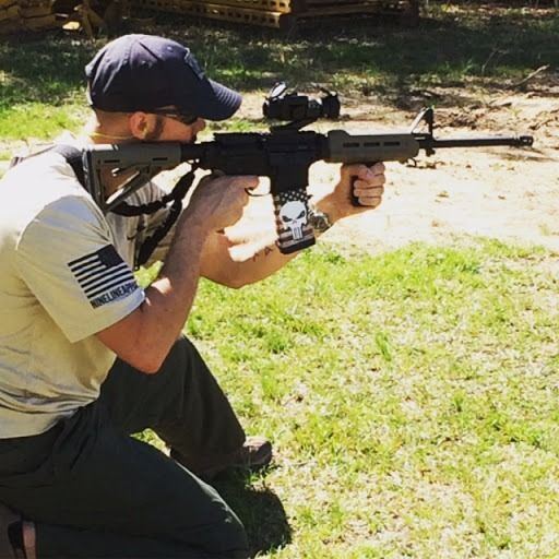 A person in a tan shirt and baseball cap kneels outdoors, aiming a rifle with a custom flag-themed magazine.