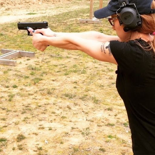 A person wearing ear protection and a black t-shirt holds a handgun with both hands at an outdoor shooting range.