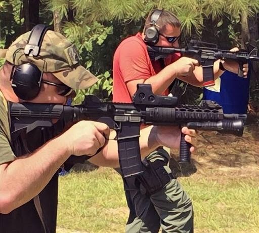 Two people wearing protective hearing gear and safety glasses practice shooting with rifles at an outdoor range.