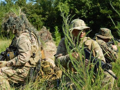 Soldiers in camouflage gear and boonie hats crouch in tall grass, holding rifles during a field training exercise.
