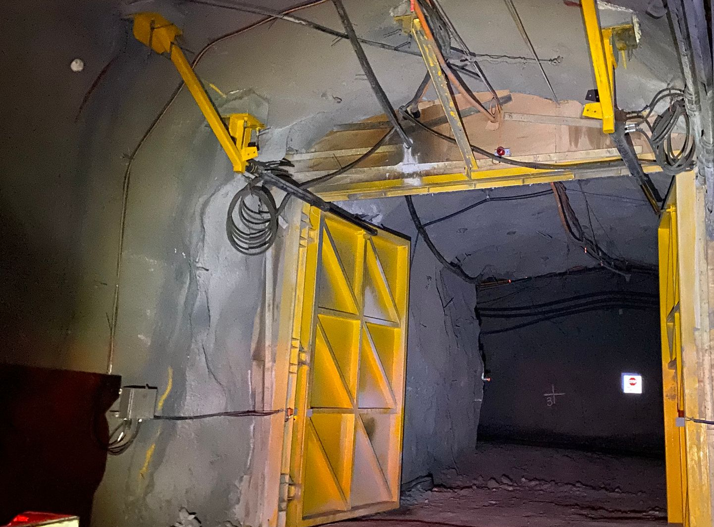 Interior of an industrial tunnel with yellow support beams and hanging cables in a dim, concrete corridor