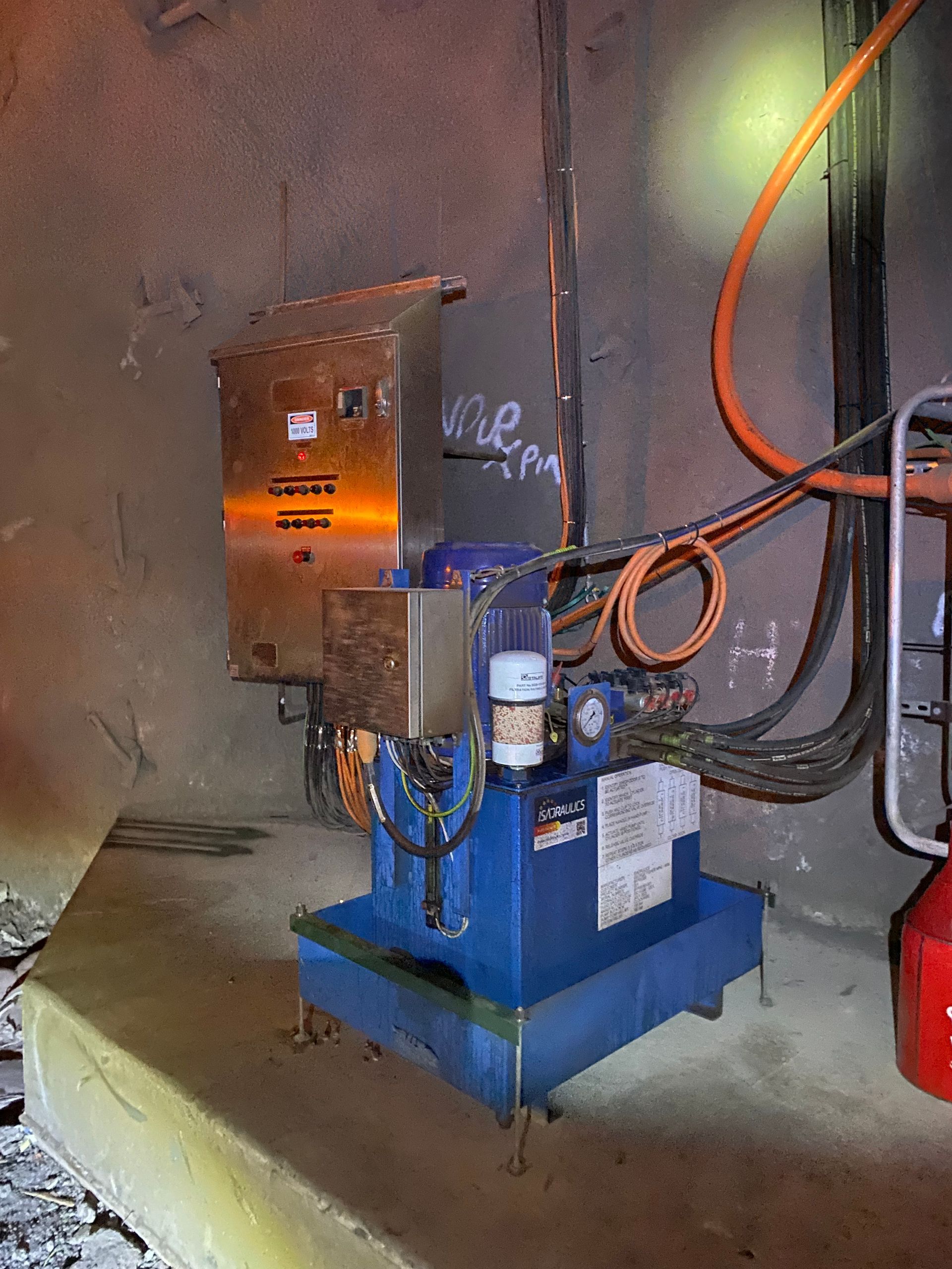 Blue industrial machine with orange housing, wires, and hoses in a concrete utility room