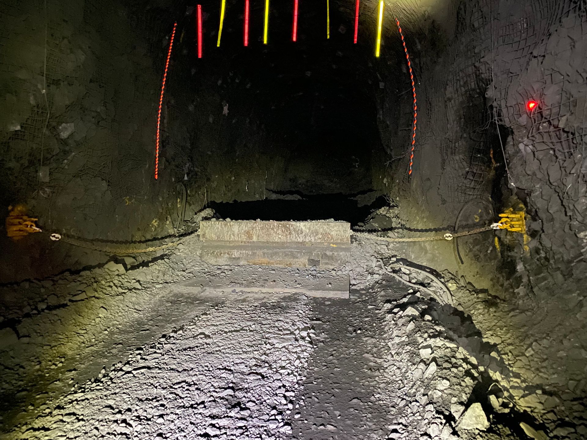 Rough tunnel interior with wet gravel road, concrete barrier, and colored survey markers overhead