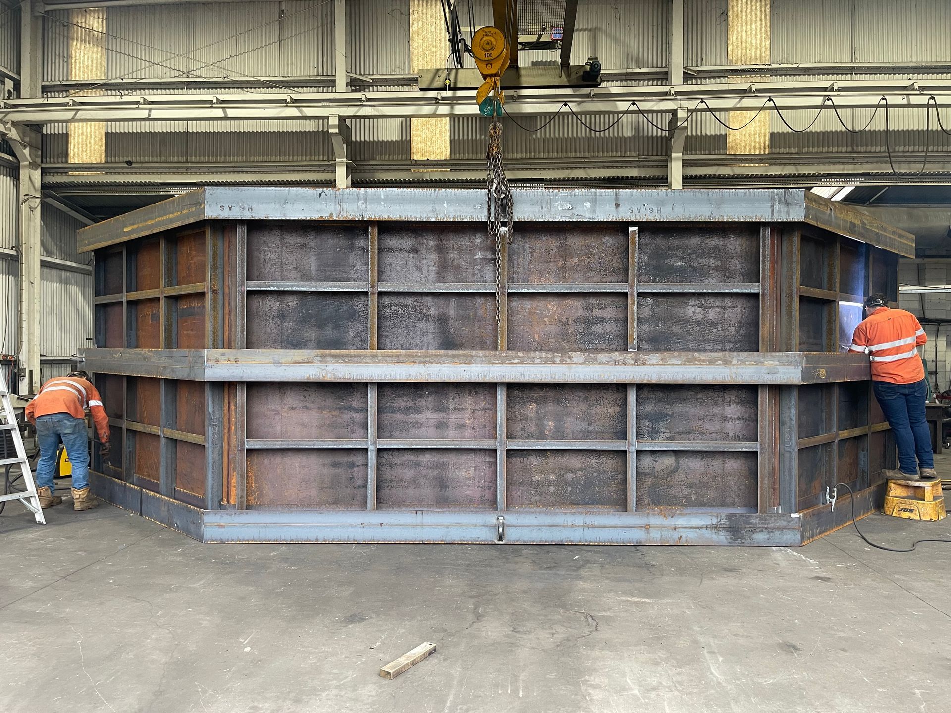 Two workers in orange vests guide a large metal industrial container inside a warehouse — Vent Doors Australia in Ryan, QLD
