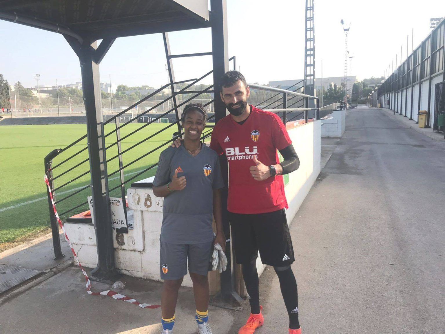 Two people posing with thumbs up beside a soccer field under a stadium walkway.