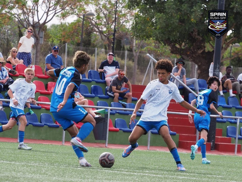 Youth soccer players in blue and white uniforms contest a ball on a turf field, with spectators in the background