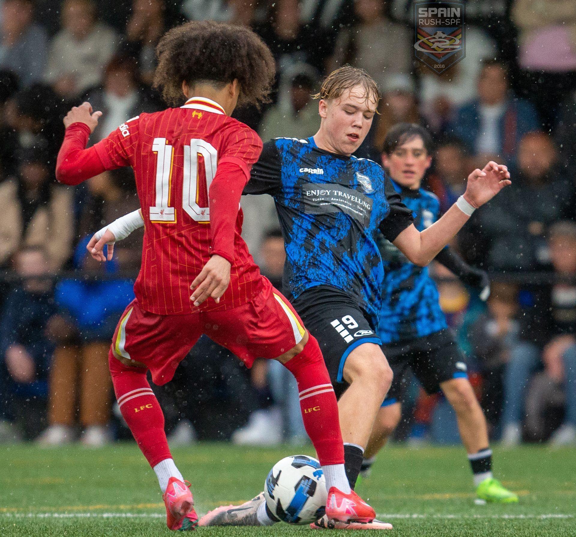 Two soccer players battle for the ball in the rain, one in red and one in blue-black stripes.