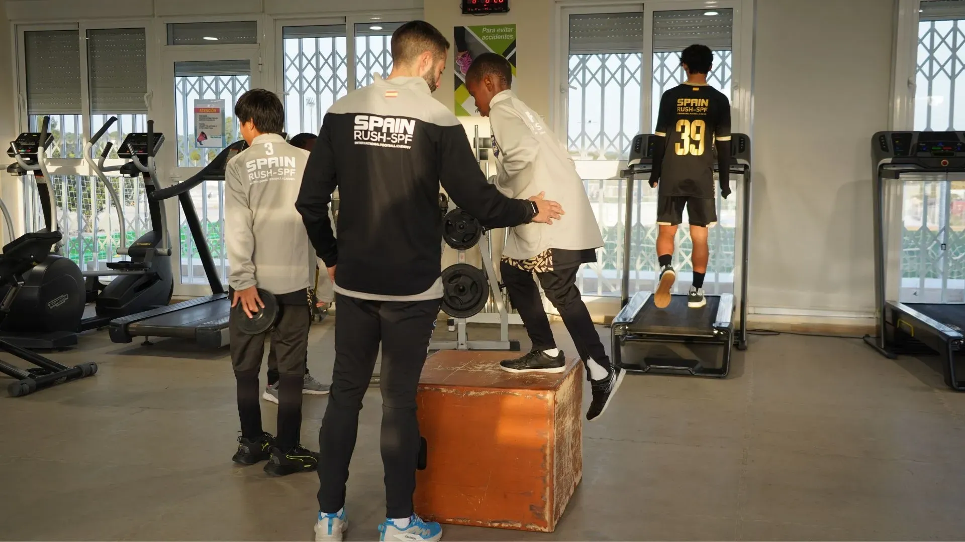 People standing in a gym near exercise equipment, with one person on a treadmill in the background.