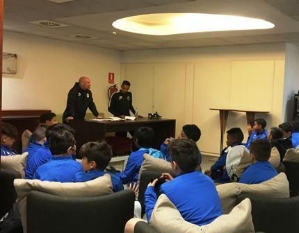 Police briefing a group of seated people in a small conference room with beige walls and overhead lighting