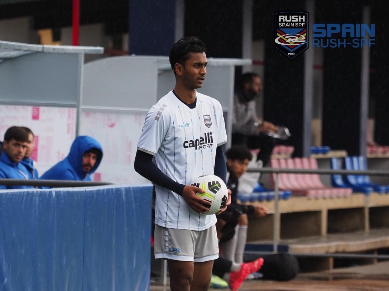 Soccer player in white kit holding a ball on the sideline, with bench and officials in the background