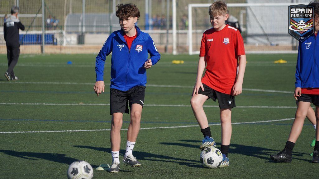 Two soccer players in blue and red jerseys standing on a field with soccer balls during practice