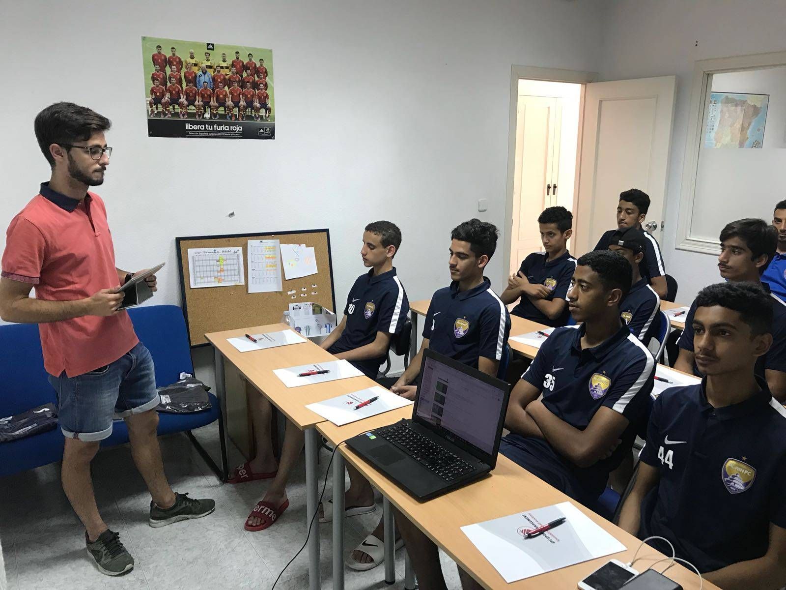 Instructor speaking to seated students in a classroom during a lesson, with laptops and notebooks on desks.