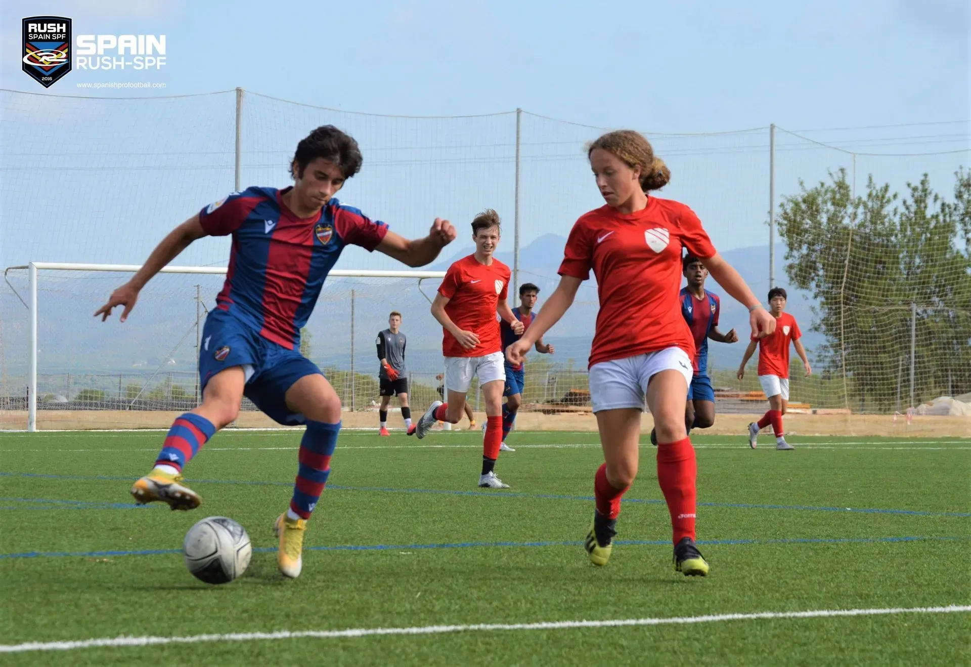 Two soccer players in red and blue jerseys chase the ball on a grassy field