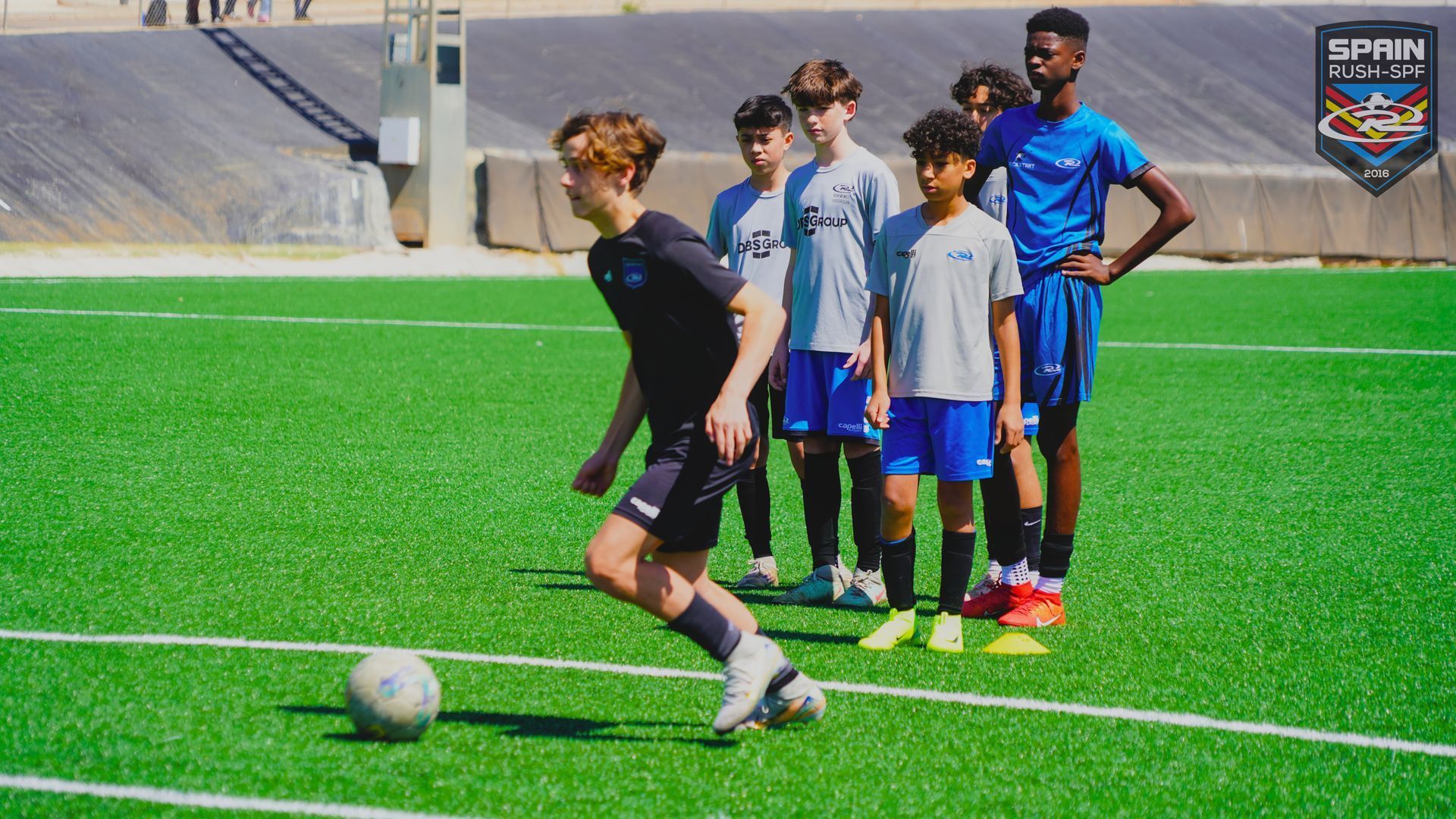 Soccer player in black prepares to kick a ball as teammates stand behind on a sunny field.