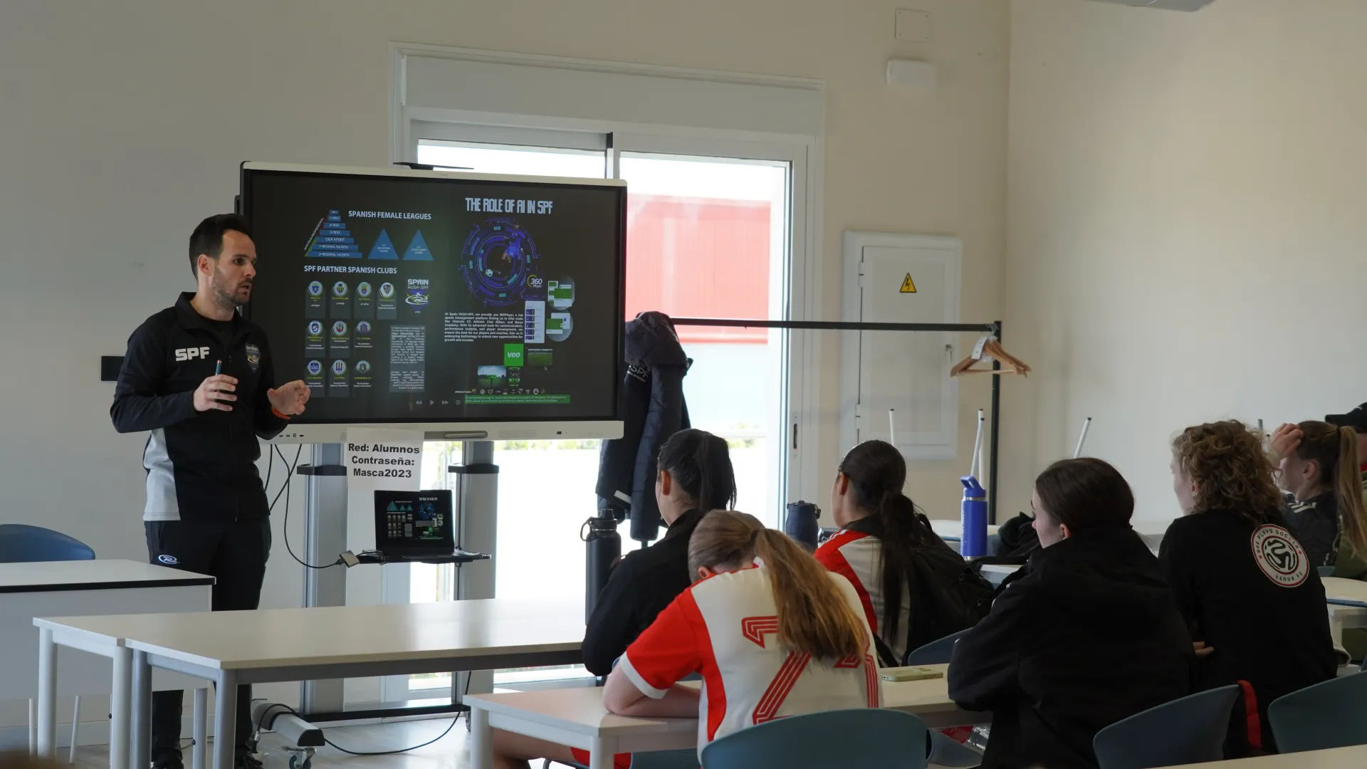 Instructor presenting to seated audience in a bright classroom with a projector screen and laptop.