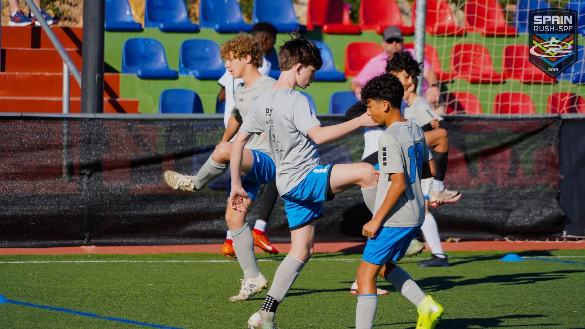 Soccer players in gray and blue uniforms contest the ball on a field with stadium seats in the background