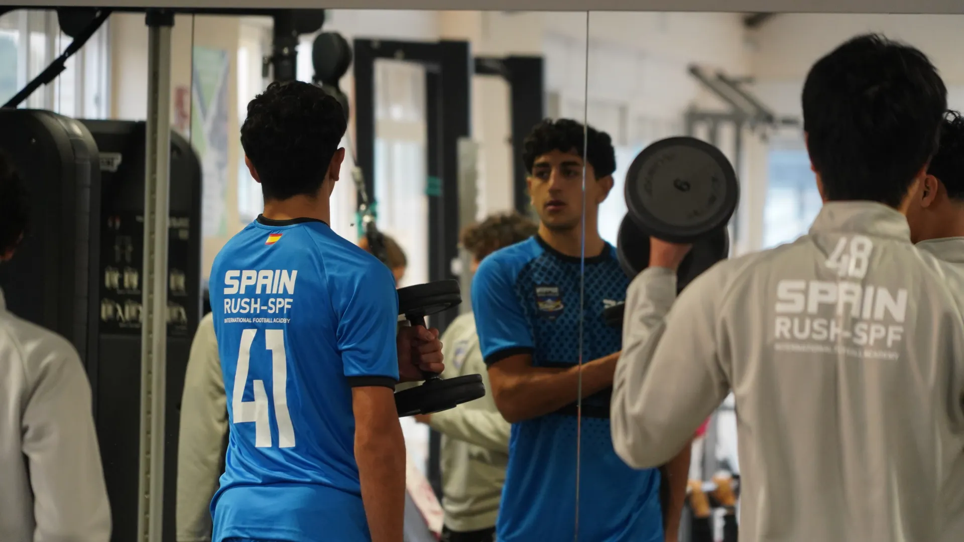 People lifting weights in a gym, seen in mirrors, with one wearing a blue shirt and another a gray shirt.