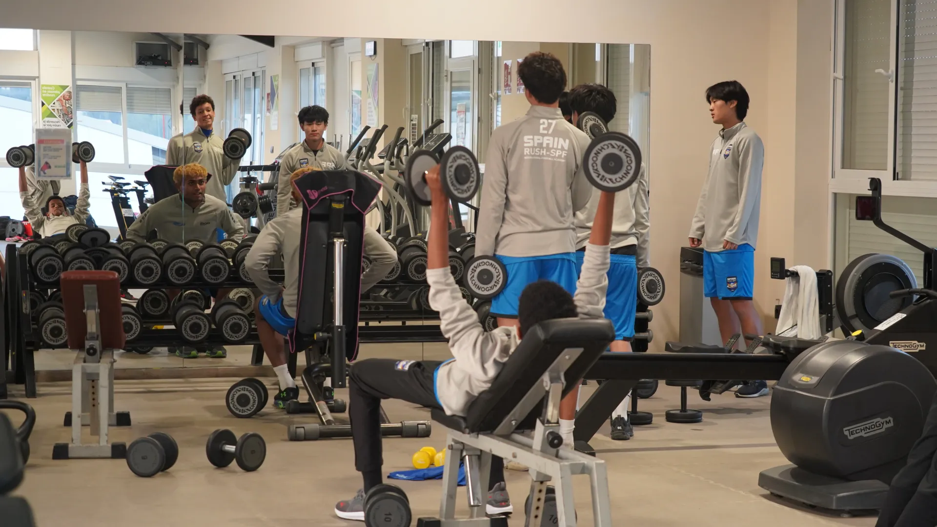 People exercising in a gym with weights, benches, and mirrors.