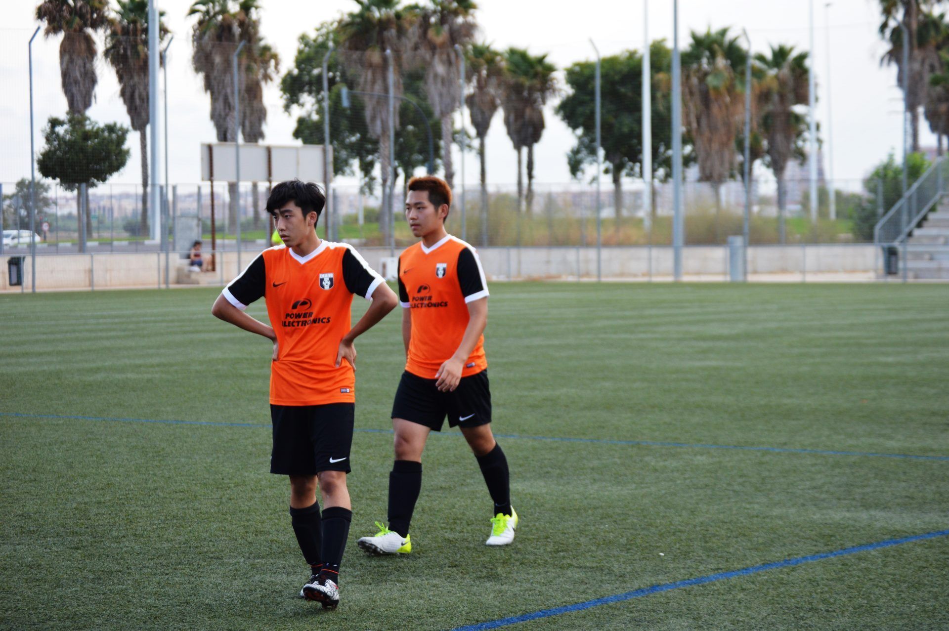 Two soccer players in orange jerseys on a field with palm trees in the background