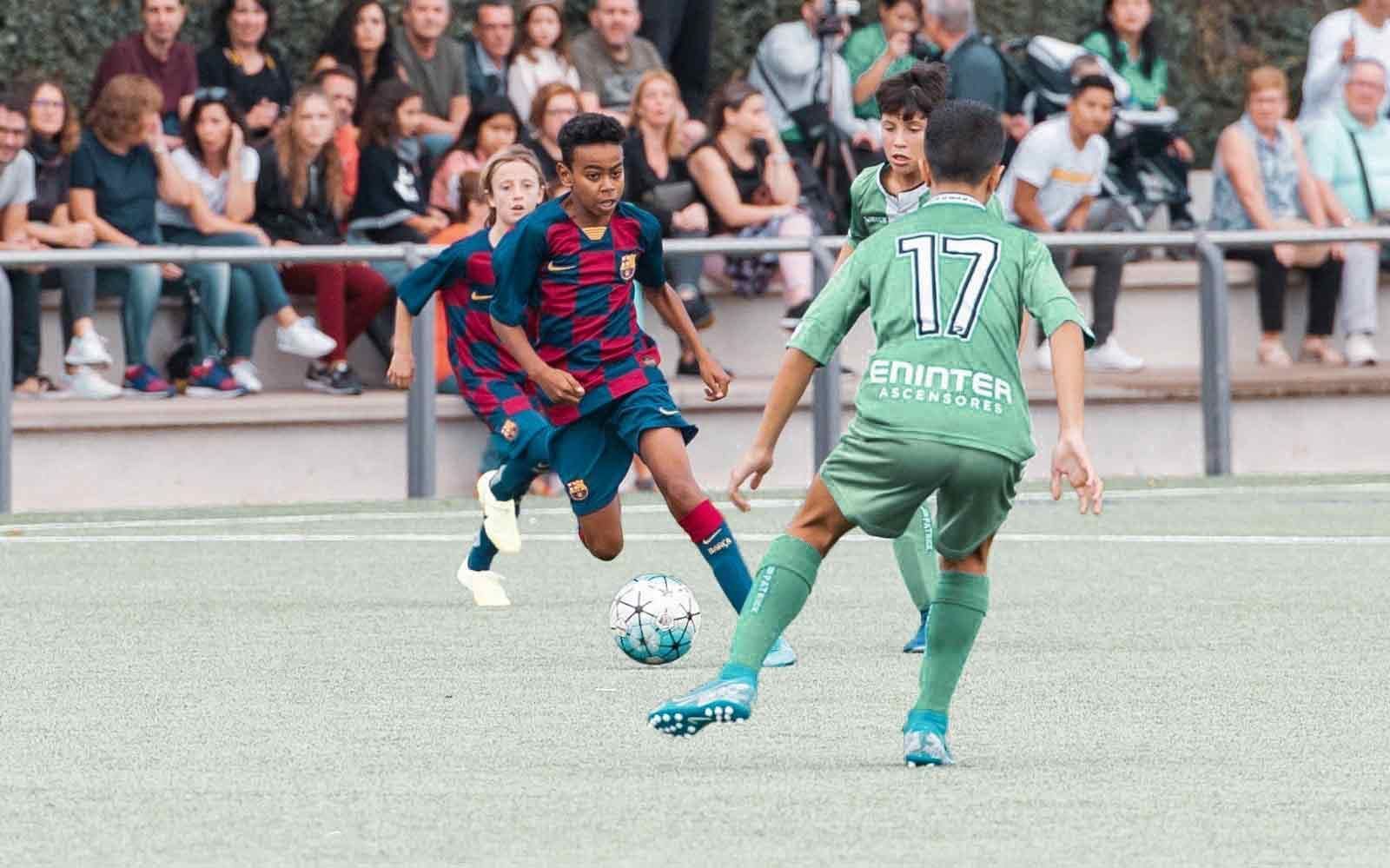 Two soccer players in red-blue and green uniforms compete for the ball on a field, with spectators behind them.