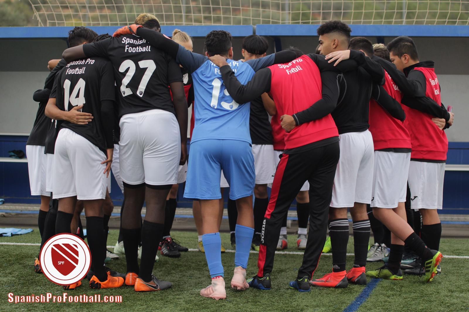 Soccer team huddled on the field, players in black, blue, and red kits with arms around each other.