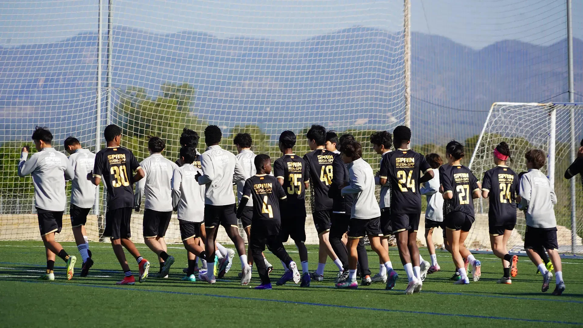 Soccer players in black and white jerseys jogging on a field near a goal, with mountains in the background