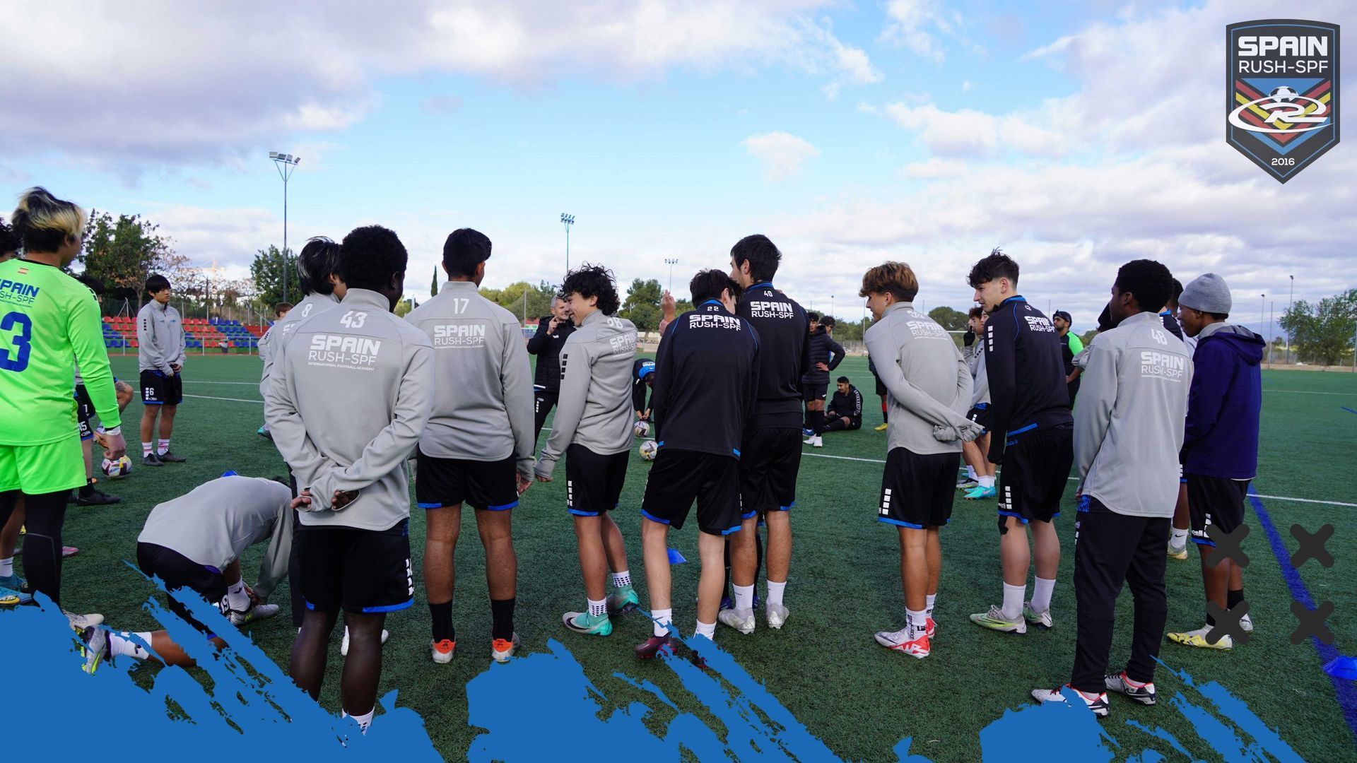 Soccer team huddled on a grassy field under a cloudy sky, viewed from behind.
