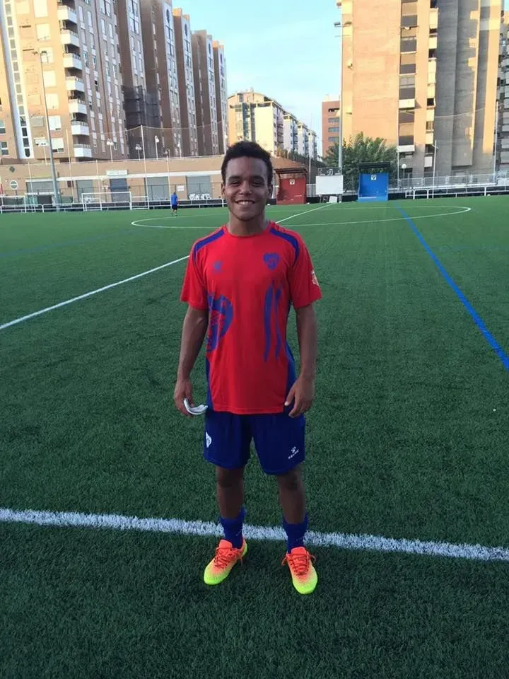 Person standing on a soccer field wearing a red jersey, blue shorts, and neon cleats.