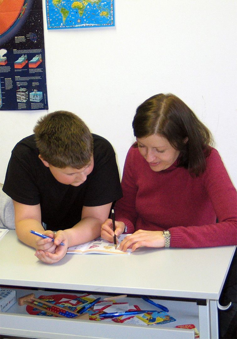 Two people leaning over a desk, writing with pencils in a classroom setting.