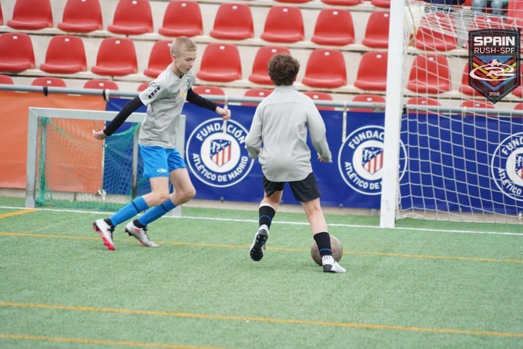Two soccer players chase the ball near the goal on an indoor field with red seats in the background