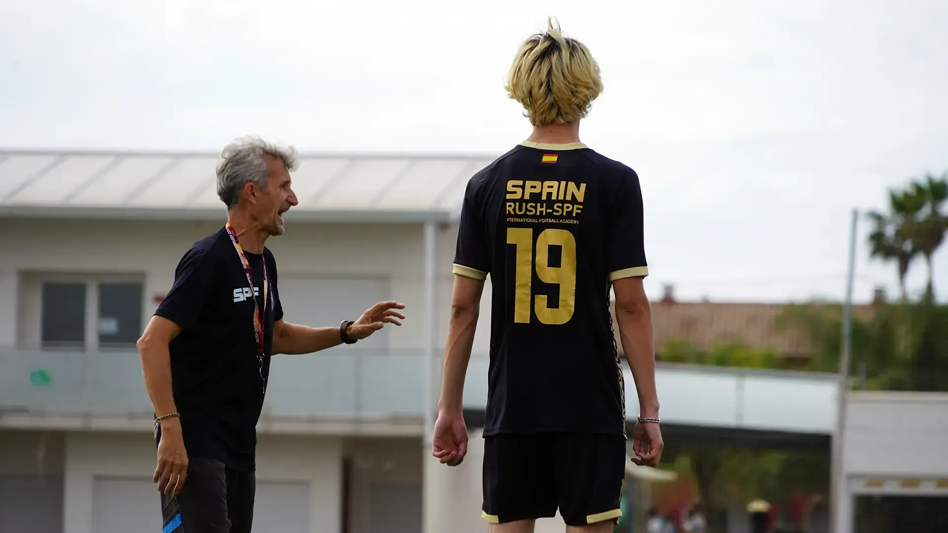 Coach speaking to a player in a black-and-gold soccer jersey on a field, with buildings in the background.
