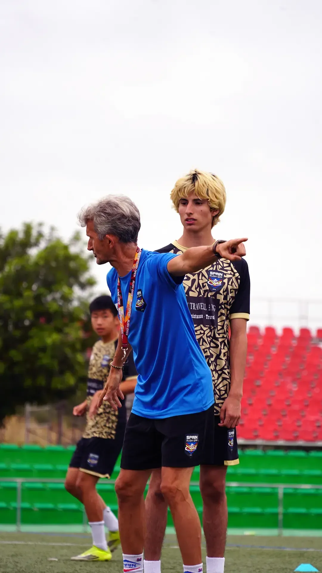 Two people standing on a sports field, one in blue pointing ahead, red stadium seats in the background