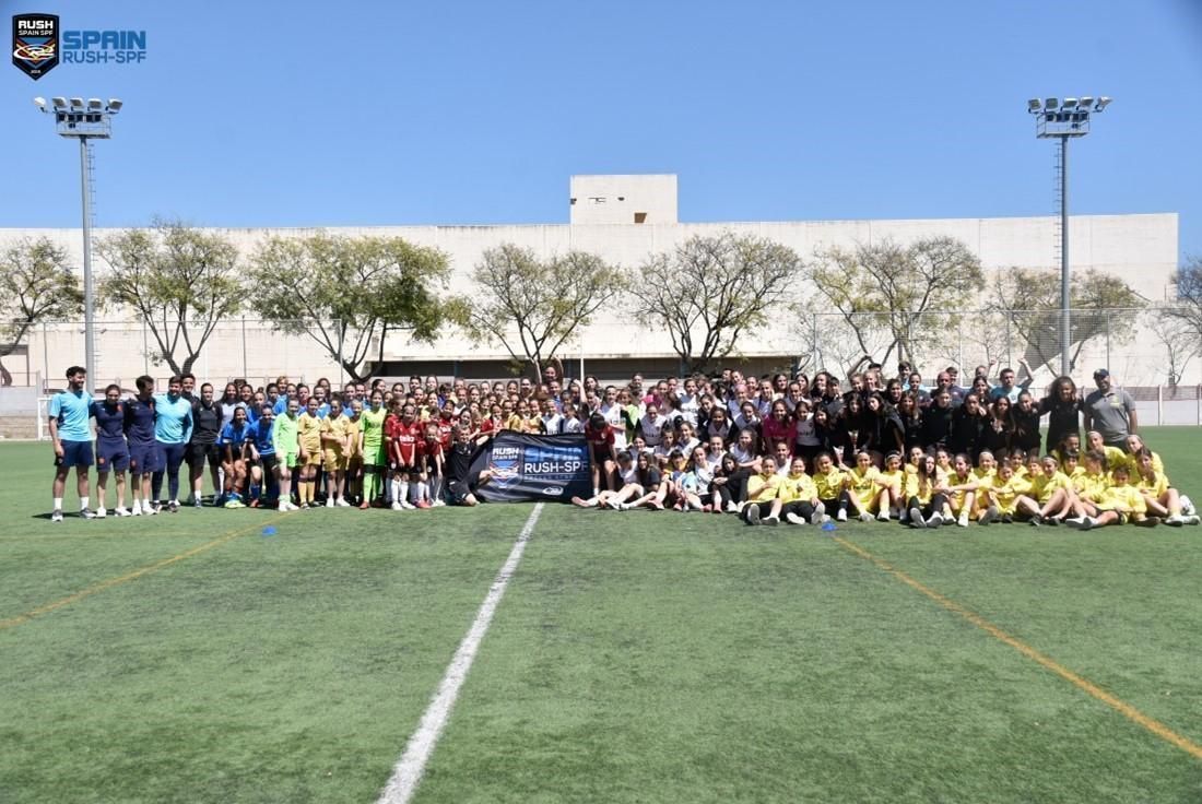 Large group posing on a soccer field with a banner, under a clear blue sky.