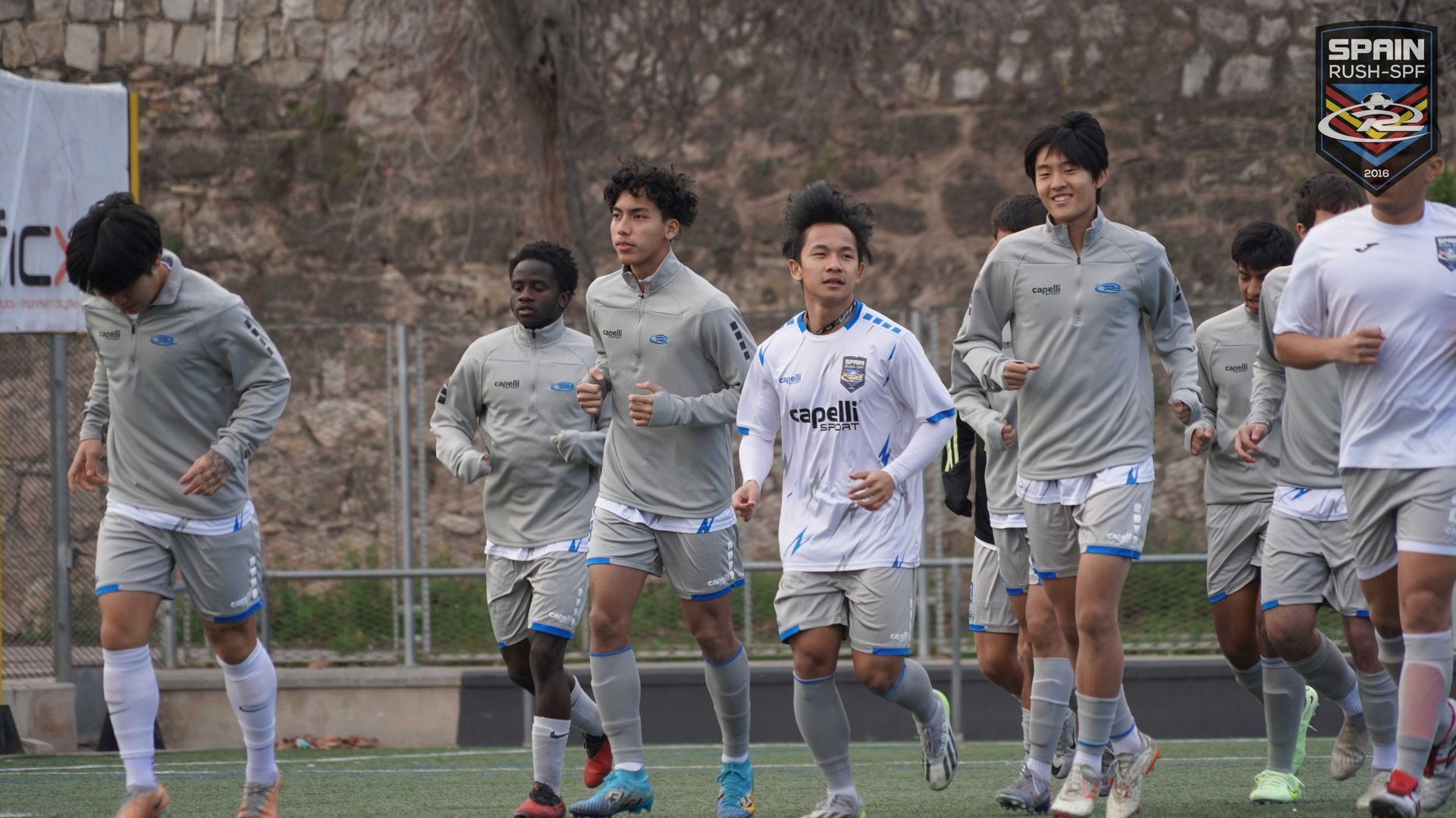 Soccer players in gray and white training gear jogging on a field near a stone wall