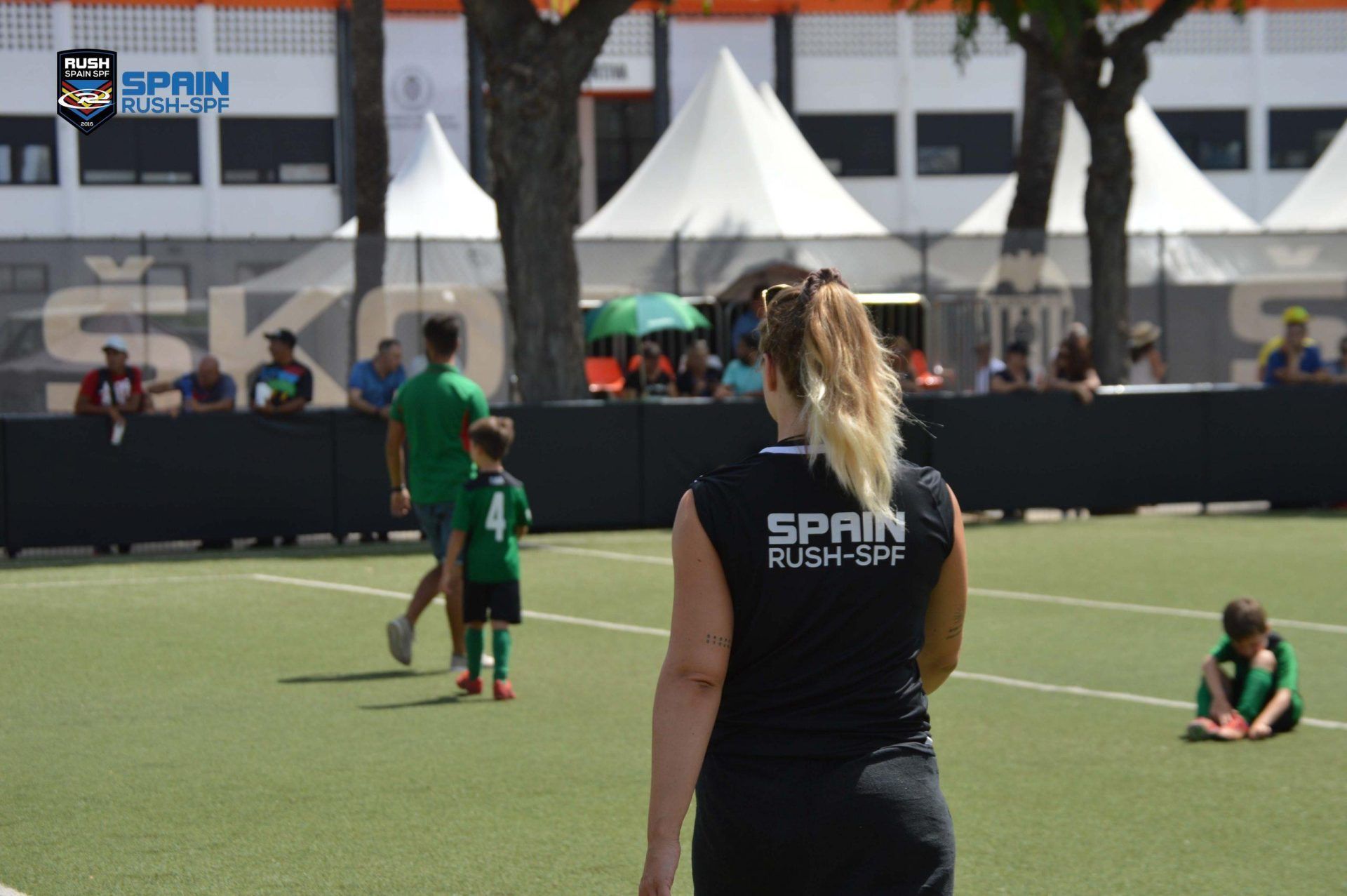 Two players in green on a soccer field, with a woman in a black shirt in the foreground and tents behind.
