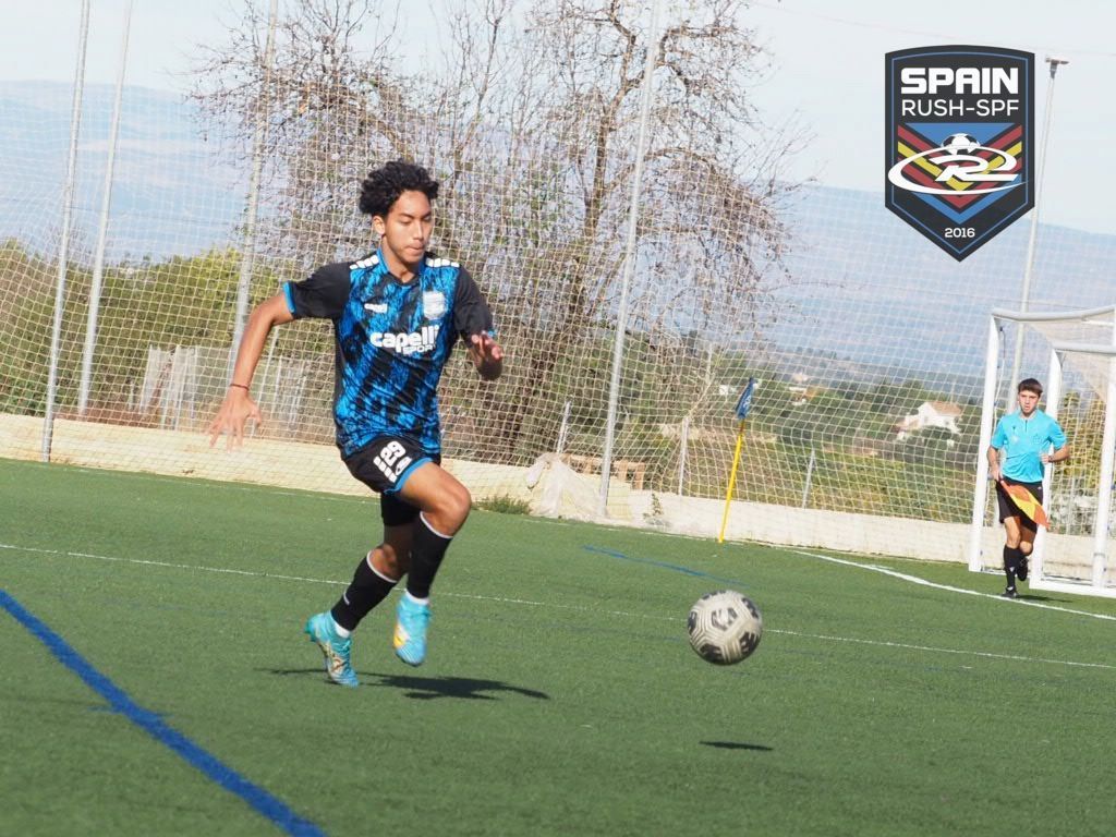 Soccer player in blue dribbles a ball on a field, with a goal and goalkeeper in the background.