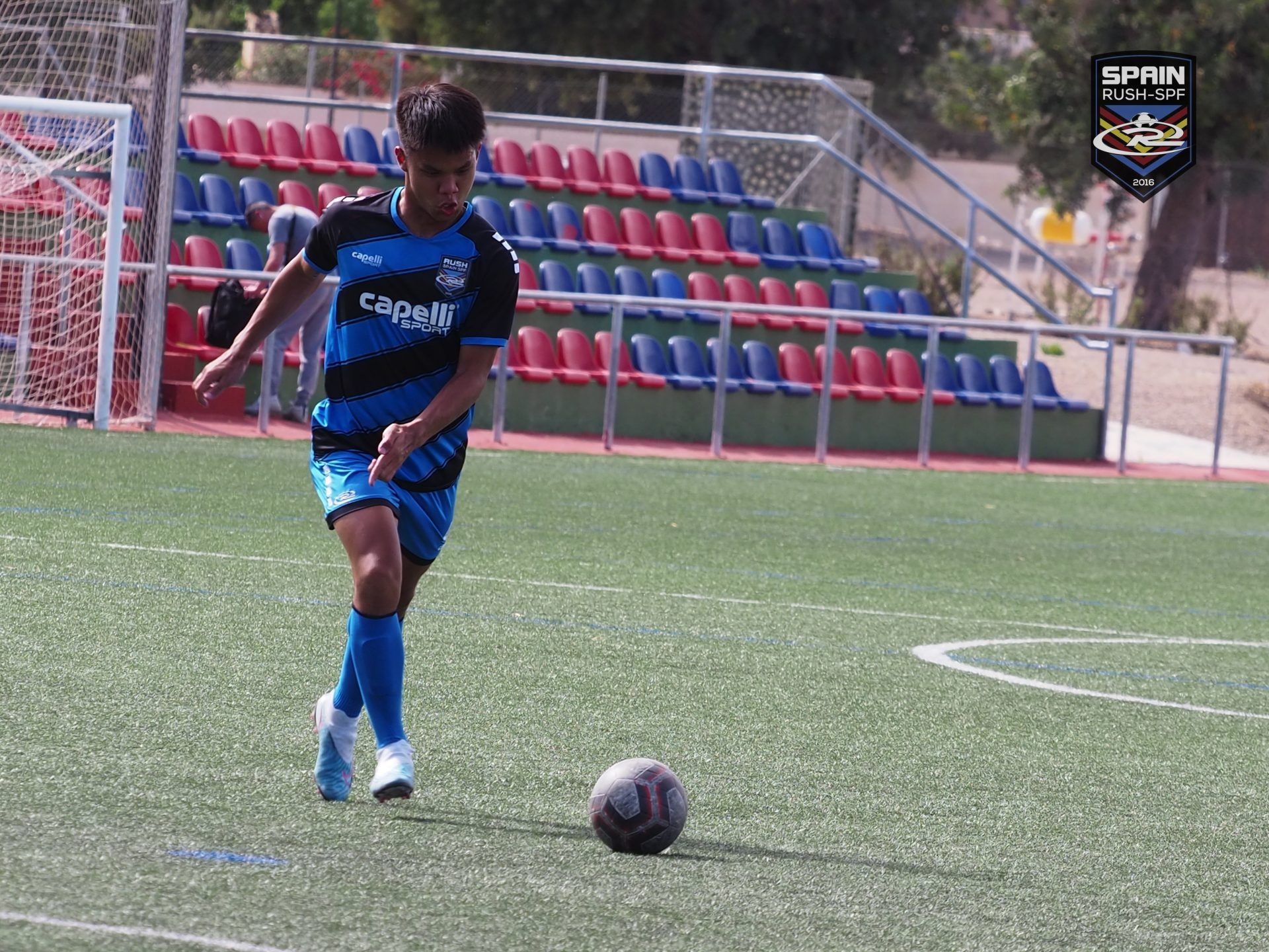 Soccer player in blue dribbles a ball on an outdoor field with red and blue bleachers behind him.