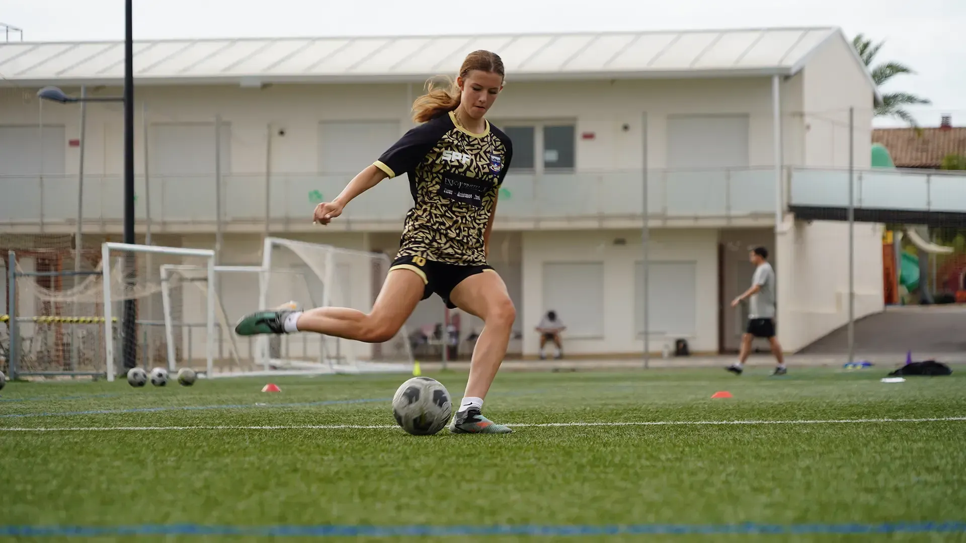 Soccer player in black and gold kit kicks a ball on a grassy field near a goal