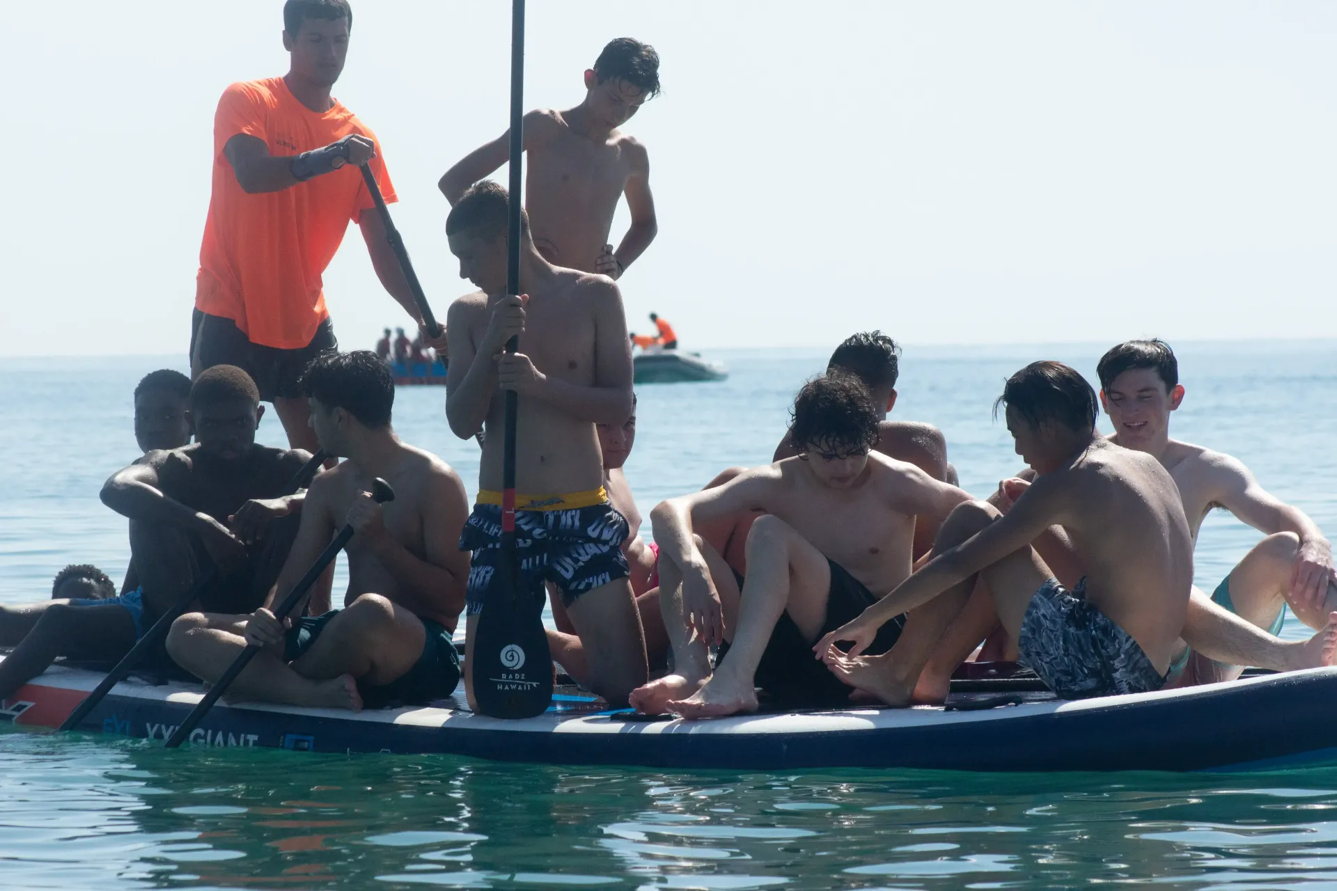 People crowded on a small boat in calm water, with one person standing in an orange shirt.